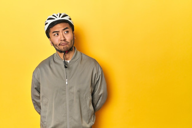 Homme asiatique en casque de vélo et gants fond de studio jaune confus se sent dubitatif et incertain