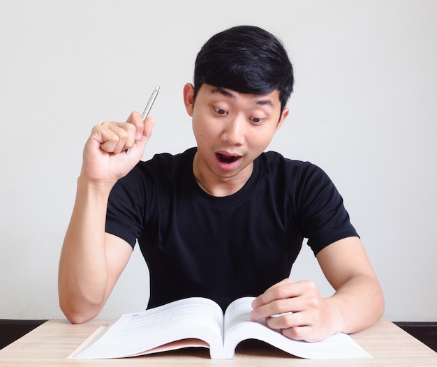 Un homme asiatique choqué et excité montre le stylo et regarde le livre sur le bureau sur blanc isolé