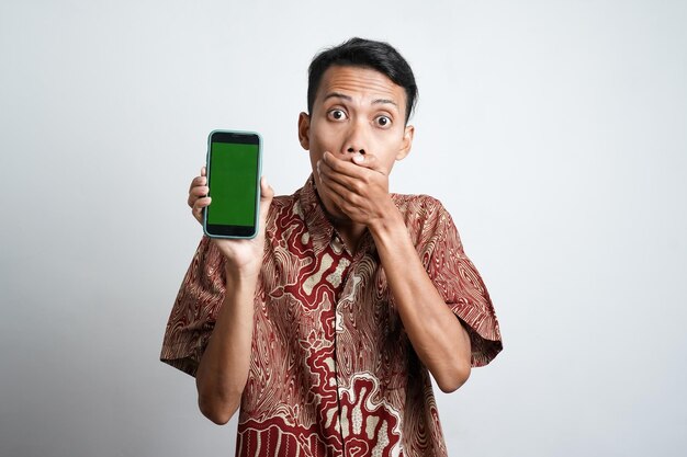 Homme asiatique choqué portant une chemise batik et montrant un écran vert sur son téléphone isolé par fond blanc