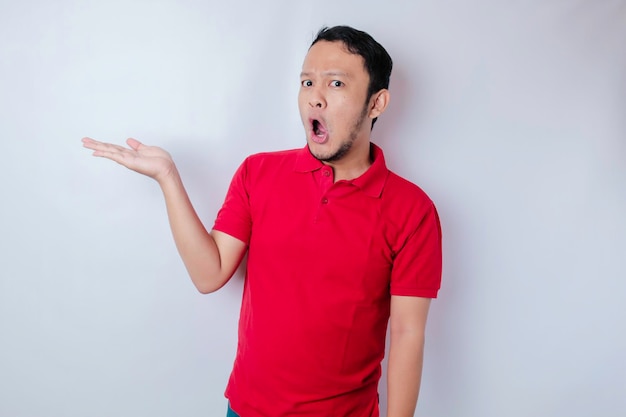 Homme asiatique choqué portant un t-shirt rouge pointant vers l'espace de copie à côté de lui isolé par un fond blanc