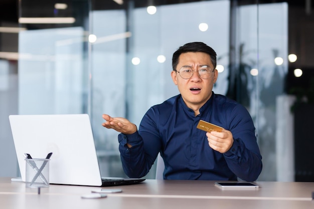 Homme asiatique en colère frustré à l'intérieur du bureau patron mature regardant la caméra et tenant la carte de crédit bancaire