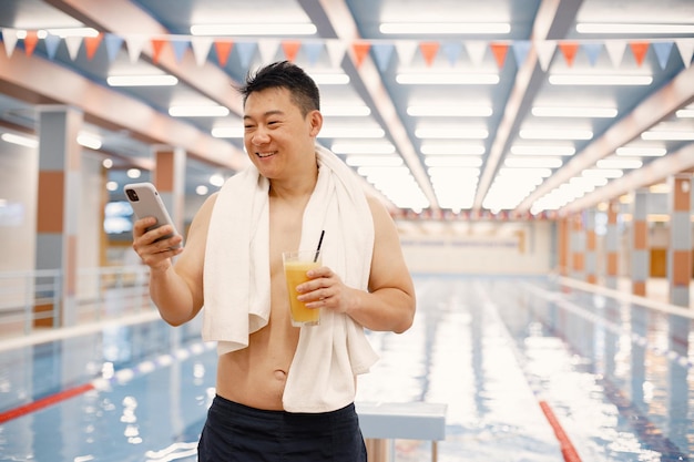 Homme asiatique debout dans une piscine intérieure et utilisant un téléphone