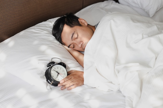 Homme asiatique dormant sur le lit sous la couverture avec réveil montrant 7 heures