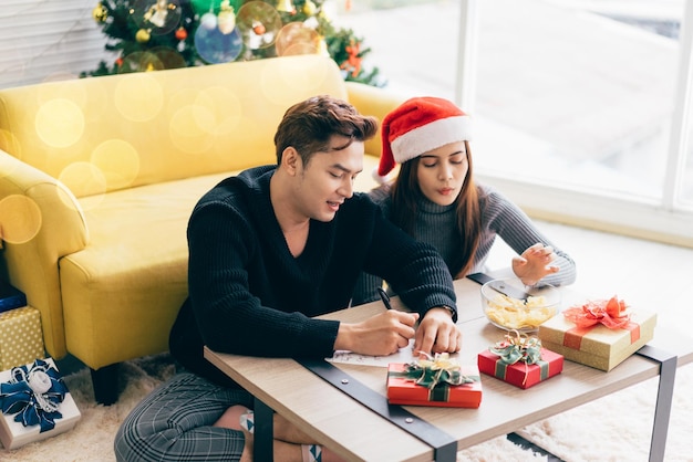 Un homme asiatique écrit sur une carte postale de Noël vierge avec un stylo avec sa petite amie Couple assis et écrivant une carte de Noël ensemble pour l'envoyer avec des cadeaux surprises à la maison pendant les vacances de Noël