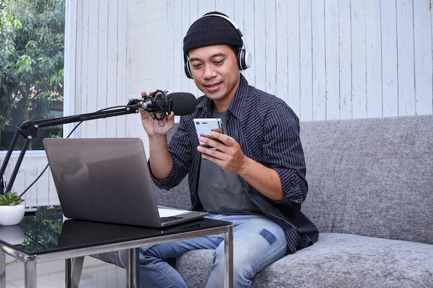 Homme asiatique enregistrant un podcast avec un casque et un microphone sur son ordinateur portable tout en tenant un téléphone portable