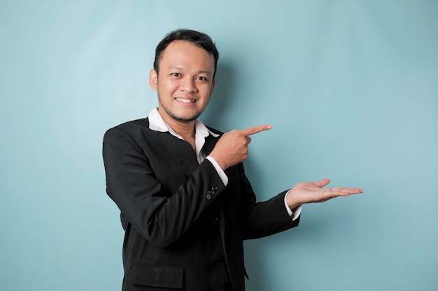 Homme asiatique excité portant un costume pointant vers l'espace de copie à côté de lui et souriant isolé par fond bleu