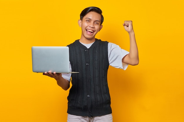 Homme asiatique excité tenant un ordinateur portable et célébrant la victoire souriant sur fond jaune