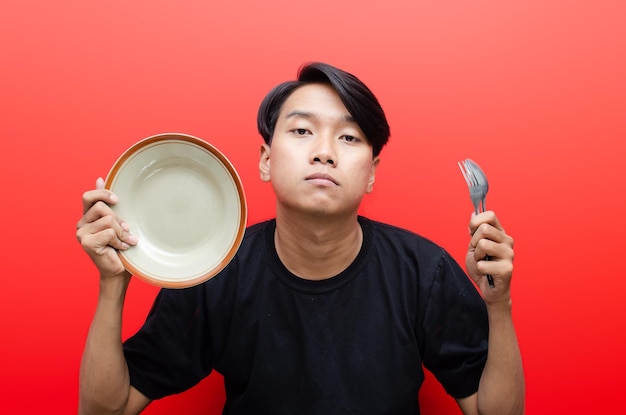 homme asiatique avec une expression triste tenant une assiette vide, une cuillère et une fourchette. Concept de jeûne et de régime