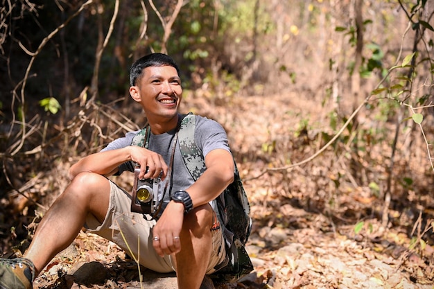 Homme asiatique heureux et actif avec équipement de trekking et son appareil photo rétro est assis sur le rocher en se reposant