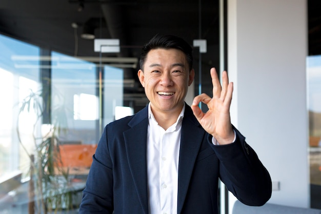 Homme asiatique heureux regardant la caméra montrant les pouces vers le haut et signe ok homme d'affaires souriant dans un bureau moderne