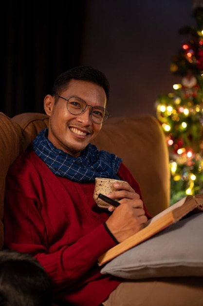 Un homme asiatique heureux se détend sur un canapé le soir de Noël en sirotant du chocolat chaud et en lisant un livre