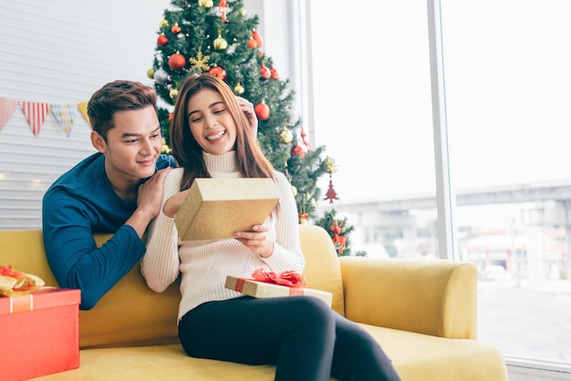 Un homme asiatique heureux surprend sa petite amie avec des cadeaux de Noël à la maison avec un arbre de Noël dans l'image d'arrière-plan avec espace de copie