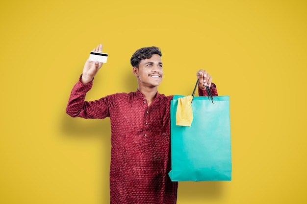 Homme asiatique indien montrant ses sacs à provisions et sa carte de crédit ou de débit tout en portant des vêtements traditionnels debout sur fond jaune