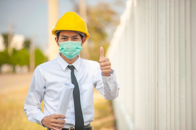 Homme asiatique ingénieur utiliser un masque facial casque jaune tenant un plan d'inspection construction construction estate