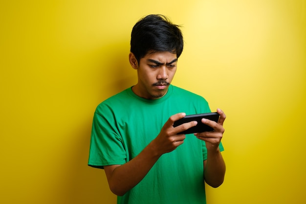 Un homme asiatique joue à un jeu mobile sur son smartphone avec une expression sérieuse ou en colère sur fond jaune
