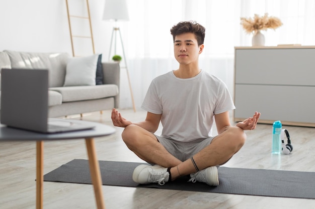 Homme asiatique méditant en position du lotus sur un tapis à l'aide d'un ordinateur