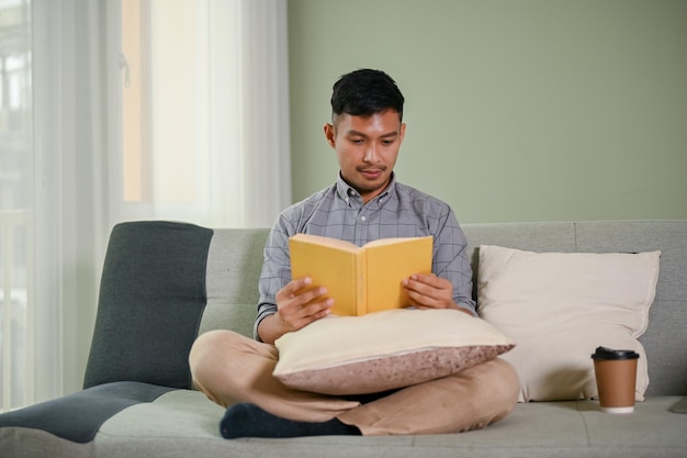 Homme asiatique millénaire intelligent et positif lisant un livre sur un canapé dans son salon