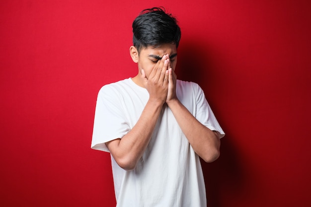 Homme asiatique avec moustache portant un t-shirt blanc avec une expression triste couvrant le visage avec les mains en pleurant. concept de dépression sur fond rouge