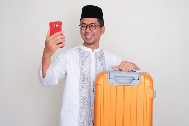 Homme asiatique musulman portant une valise montrant une expression heureuse en regardant son téléphone