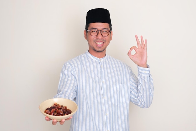 Photo homme asiatique musulman souriant et donnant un signe de doigt ok tout en tenant un bol de dattes et de fruits
