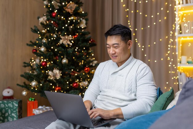 Homme asiatique à noël travaillant sur un ordinateur portable assis à la maison sur un canapé dans le salon près de l'arbre de noël