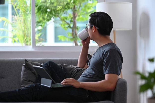 Homme asiatique paisible buvant du café et regardant par la fenêtre assis dans le salon