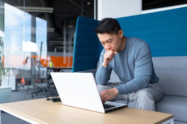 Un homme asiatique pensant travaille sur un ordinateur dans un bureau moderne un pigiste se concentre sur la résolution d'une tâche fait un travail important