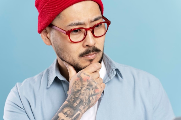 Homme asiatique pensif portant des lunettes élégantes chapeau rouge isolé sur fond bleu