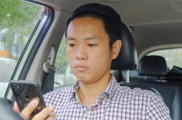 Homme asiatique portant une chemise à carreaux se penche sur un téléphone intelligent dans une voiture.