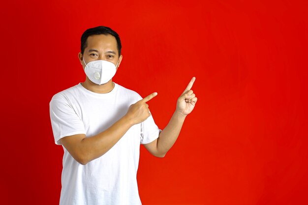 Homme asiatique portant un masque médical pointant vers le côté vide avec les deux mains sur fond rouge