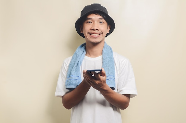 Homme asiatique portant un t-shirt blanc brillant dans un chapeau noir avec une serviette bleue portant un téléphone sur fond isolé