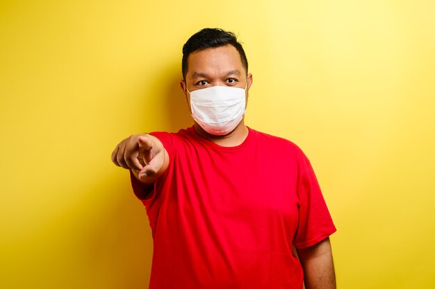 Homme asiatique portant un t-shirt rouge et un masque pointant vers l'avant, regardant la caméra en choisissant quelqu'un pour porter un masque sur fond bleu.