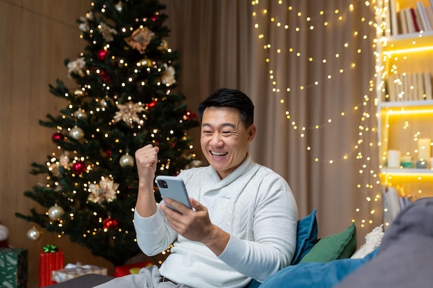 Homme asiatique satisfait à la maison près de l'arbre de Noël à l'aide de téléphone célébrant le succès et la victoire