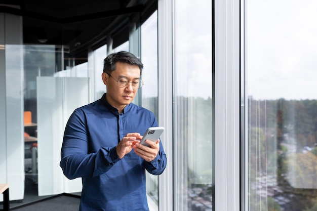 Un homme asiatique sérieux et concentré à l'intérieur du bureau utilise le téléphone, un homme d'affaires se tient debout