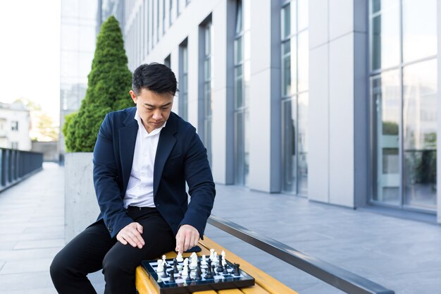 Homme asiatique sérieux jouant aux échecs à l'extérieur, homme d'affaires pensant jouer aux échecs assis sur un banc près du centre de bureaux