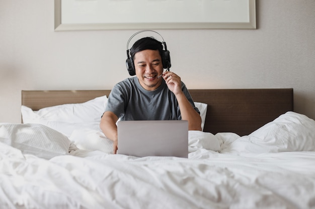 Homme asiatique souriant dans des écouteurs faisant une vidéoconférence avec un ordinateur portable sur le lit
