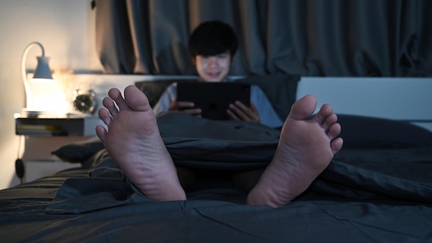 Homme asiatique souriant utilisant une tablette numérique la nuit dans la chambre.