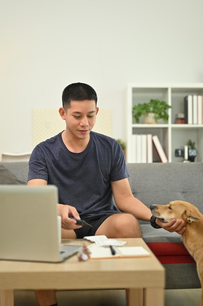 Un homme asiatique souriant en vêtements décontractés jouant avec un adorable chien alors qu'il est assis sur le canapé du salon