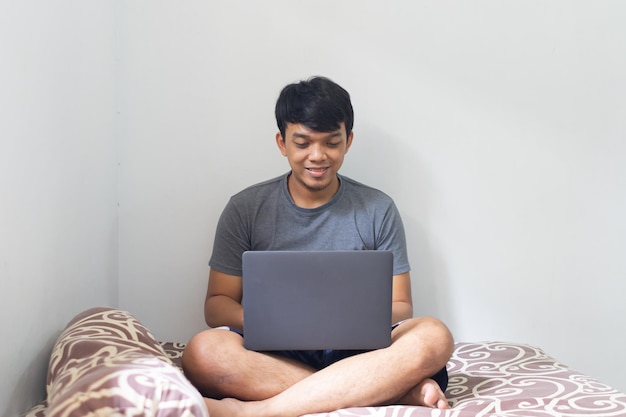 Photo un homme asiatique sourit et regarde un ordinateur portable dans une chambre pour le concept de travail à domicile avec lumière naturelle