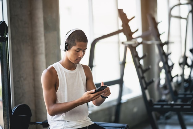 Homme asiatique sportif corps en forme portant un casque blanc et utilisant un téléphone portable pour écouter de la musique tout en se reposant ou avant de courir faire de l'exercice à la salle de fitness Sport course saine concept de salle de fitness