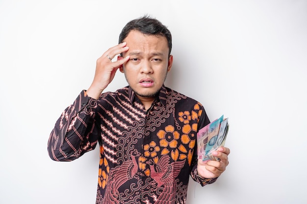Un homme asiatique stressé portant une chemise batik et tenant de l'argent en roupie indonésienne isolé par fond blanc a l'air déprimé