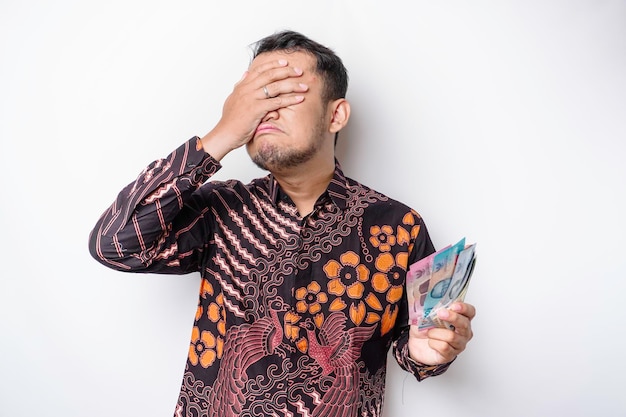 Un homme asiatique stressé portant une chemise batik et tenant de l'argent en roupie indonésienne isolé par fond blanc a l'air déprimé