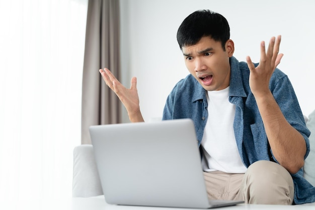 Un homme asiatique surpris choqué regardant un écran d'ordinateur portable assis dans le salon