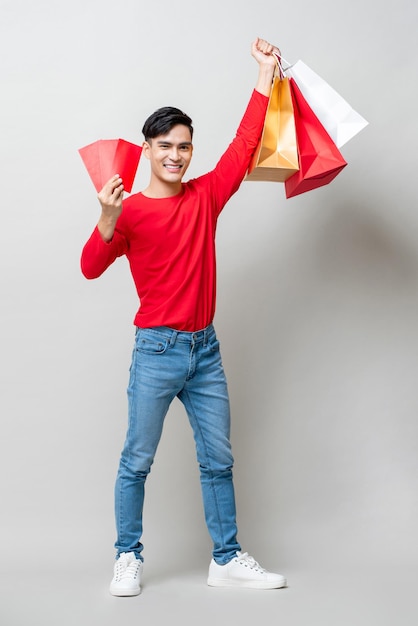 Homme asiatique surpris levant les mains montrant des sacs à provisions et une enveloppe rouge Ang Pow dans un fond de studio gris clair isolé pour le concept de vente du nouvel an chinois