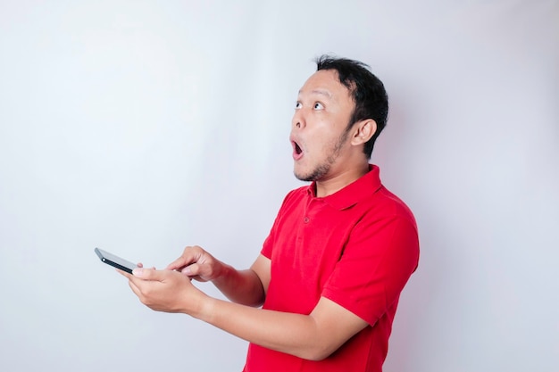 Homme asiatique surpris portant un t-shirt rouge pointant vers son smartphone isolé sur fond blanc