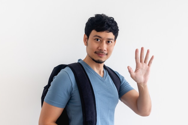 Homme asiatique en t-shirt bleu avec sac à dos agite la main sur isolé