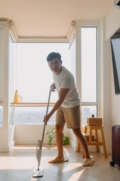 Un homme asiatique thaïlandais utilise un balai pour nettoyer le sol dans le salon de l'appartement. Un homme fait les tâches ménagères.