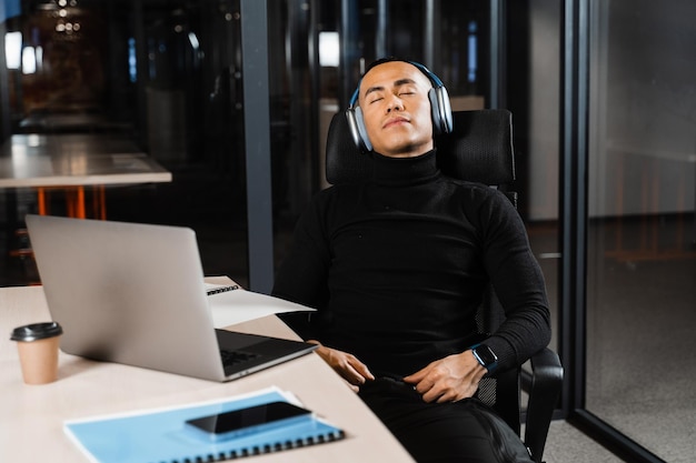 Un homme asiatique travaillant en heures supplémentaires avec les yeux fermés se repose au travail sur un ordinateur portable Un programmeur masculin fatigué prend une pause en raison d'heures supplémentaires