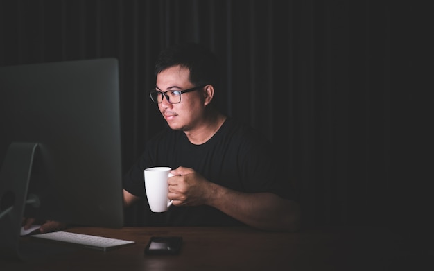 Homme asiatique travaillant sur ordinateur dans la pièce sombre et tenant une tasse de café