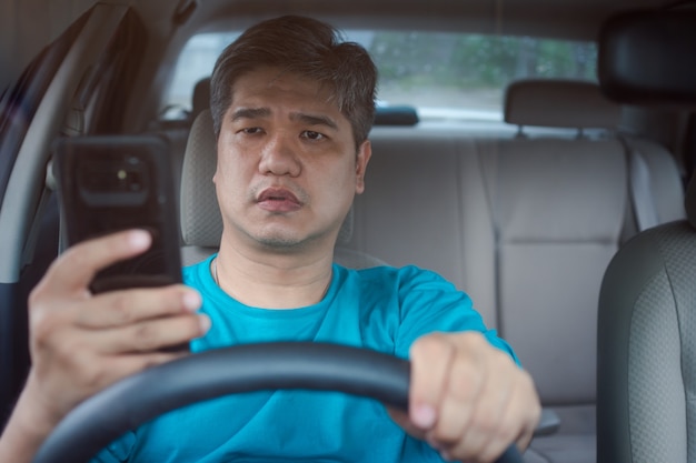 Homme asiatique utilisant un téléphone intelligent mobile et lisant des messages en conduisant une voiture. Notion de négligence, de comportement dangereux et de risque d'accident